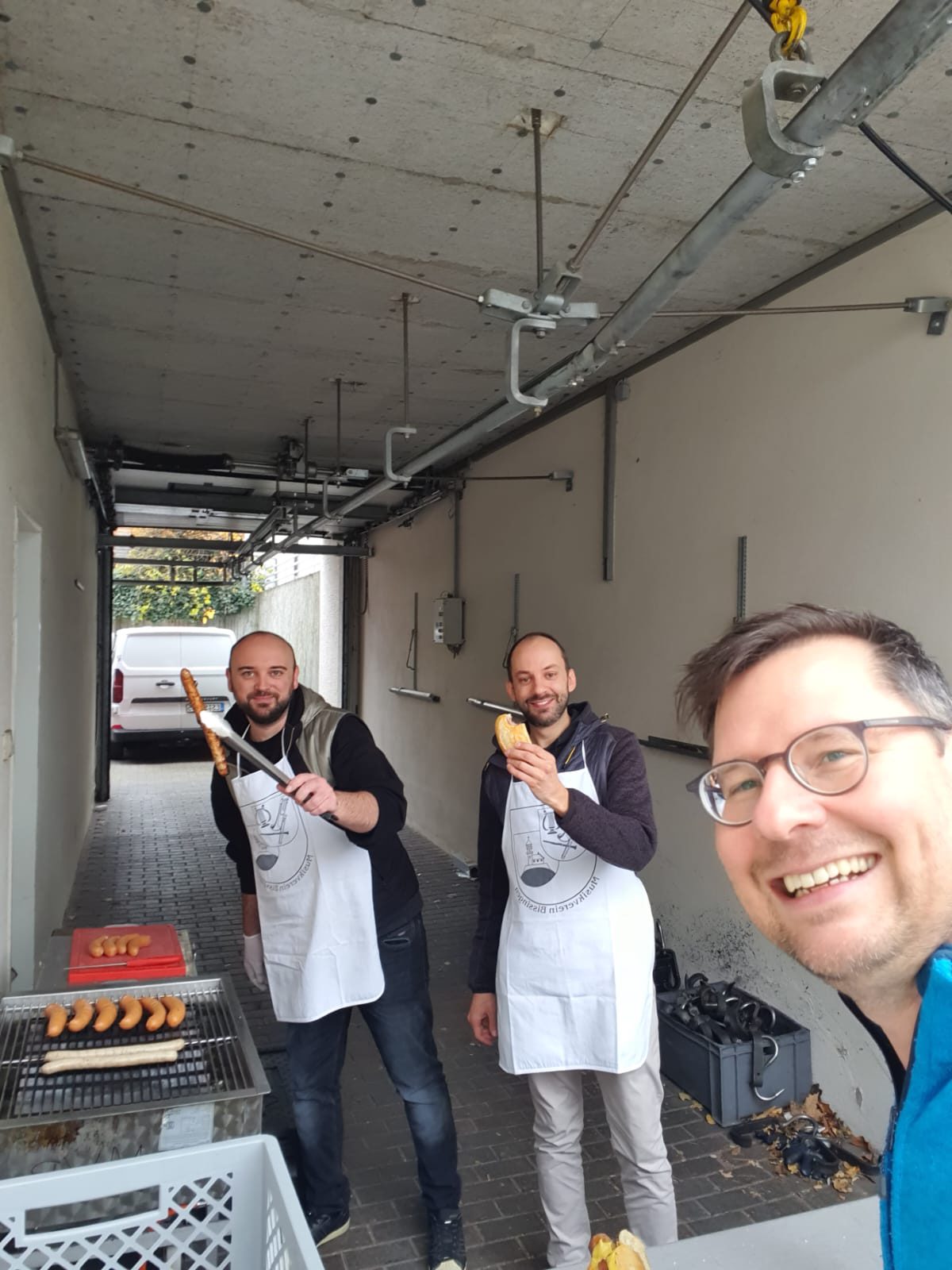 Drei Musiker in Schürzen mit Vereinswappen am Grill, einer hält eine Thüringer mit der Grillzange hoch