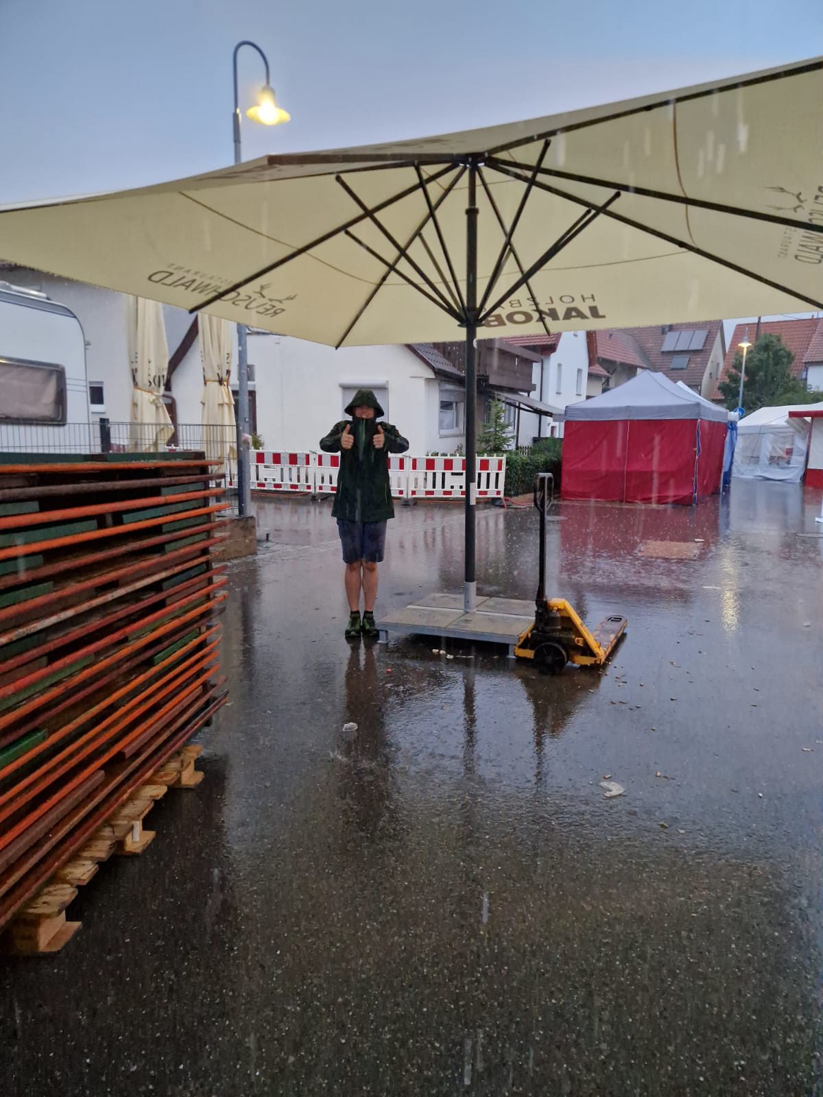 Im strömenden Regen hebt ein Helfer in Regenjacke unter einem Sonnenschirm beide Daumen, ringsum gestapelte Biertische