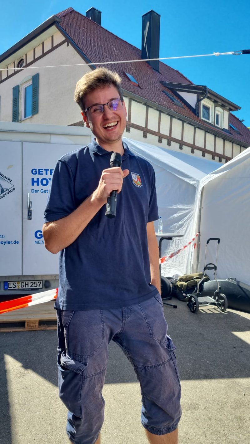 Der 2. Vorstand lacht mit einem Funkmikrofon in der Hand, im Hintergrund das Festzelt