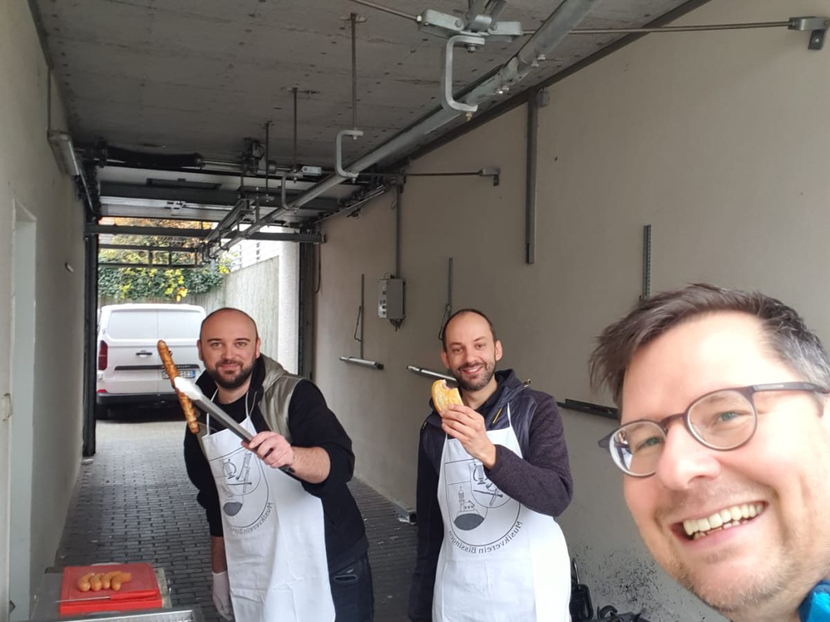 Drei Musiker in Schürzen mit Vereinswappen am Grill, einer hält eine Thüringer mit der Grillzange hoch